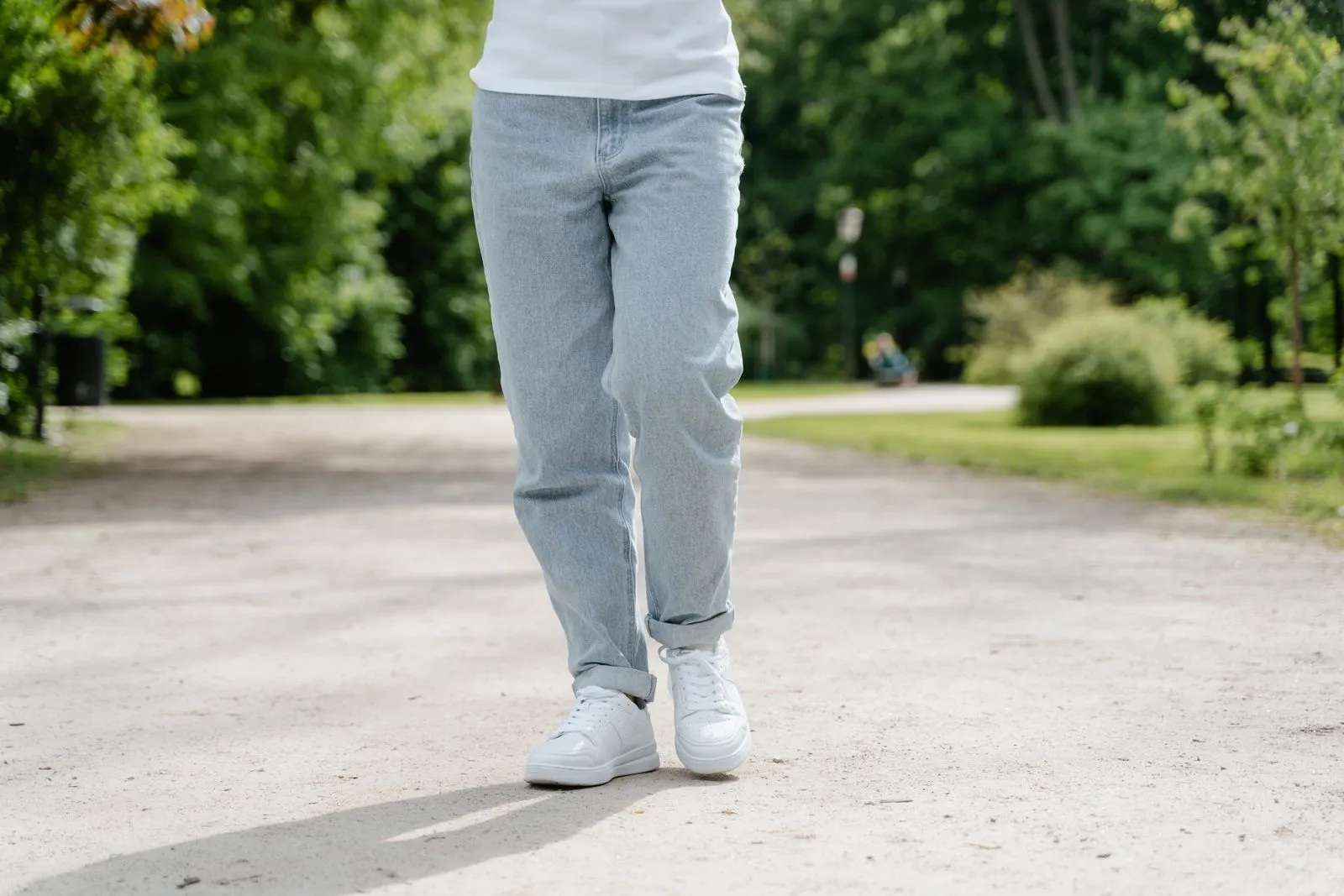 A close-up view of a person walking on a park pathway wearing blue jeans and white sneakers.